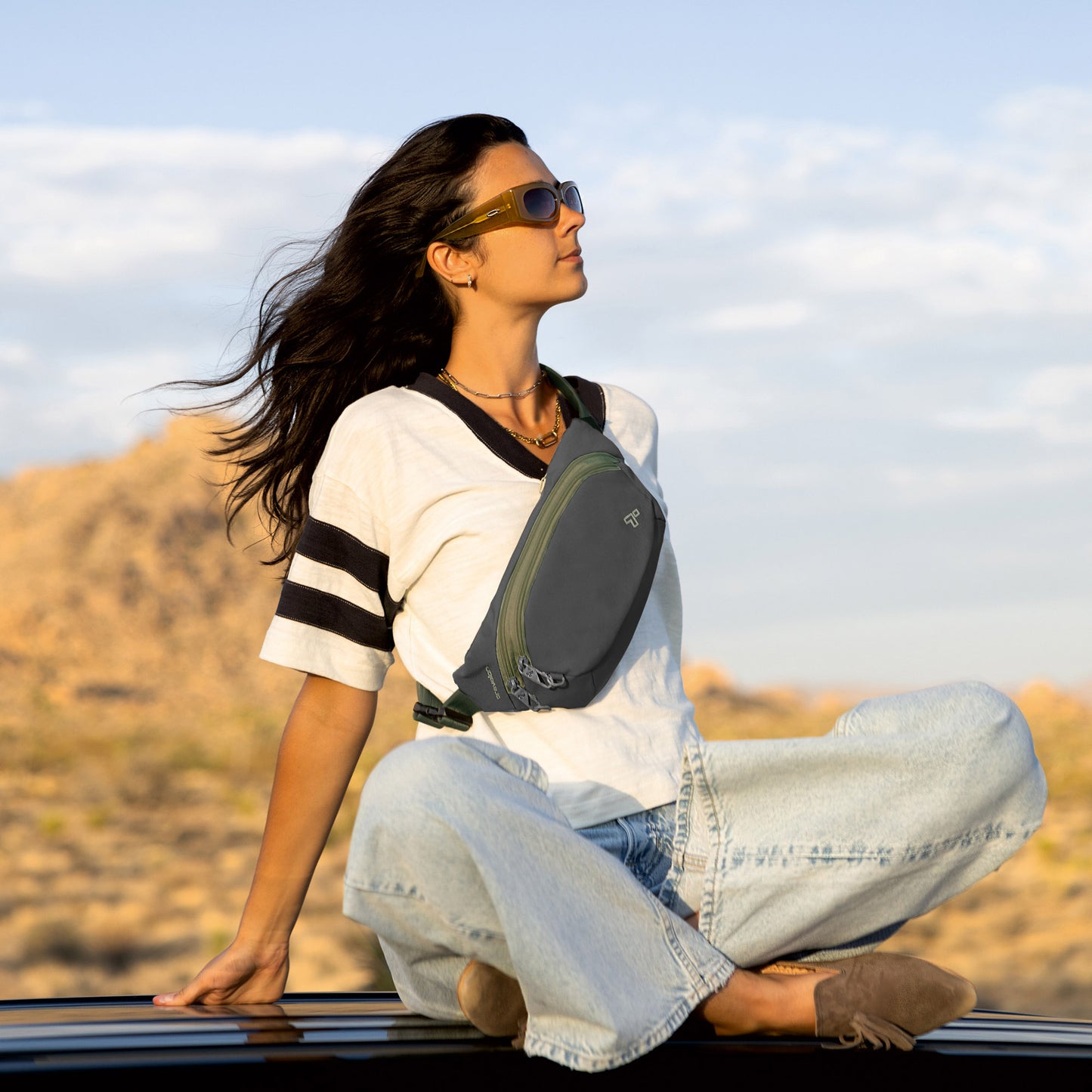Woman sits cross-legged on a car roof in the desert, wearing a Travelon Voyages Anti-Theft Waist Pack and sunglasses.