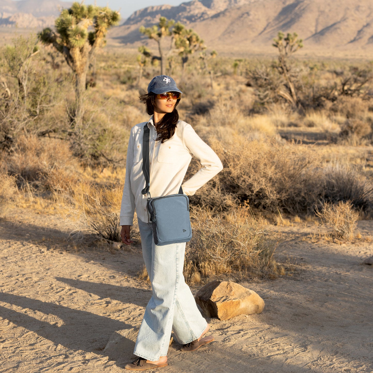 Person in a desert landscape wearing sunglasses, a hat, and the Travelon Voyages Anti-Theft Slim Bag hands-free.