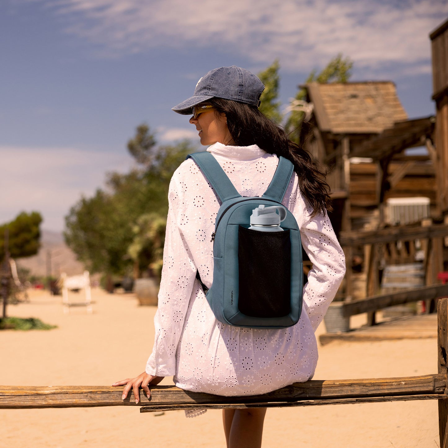 A white-dressed person in a blue cap leans on a wooden fence, sporting a Travelon Voyages Anti-Theft Backpack.