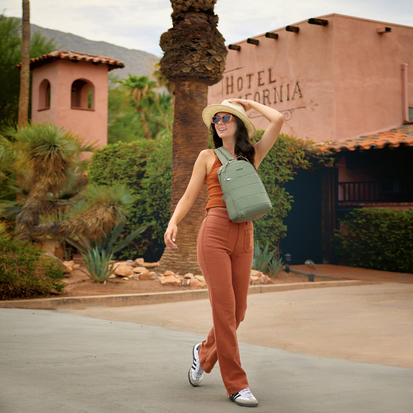 Person in rust outfit and hat walks outside with Travelon Roam Anti-theft Convertible Backpack, palm trees behind.