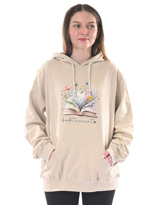 Product Image – Woman in a beige Brief Insanity Booktrovert Hoodie with an open book and flowers, hands in pockets.