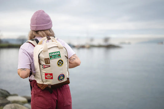 Product Image – Person with a beanie and Northern Icons Canada Everyday Backpack stands by calm water on a cloudy day.