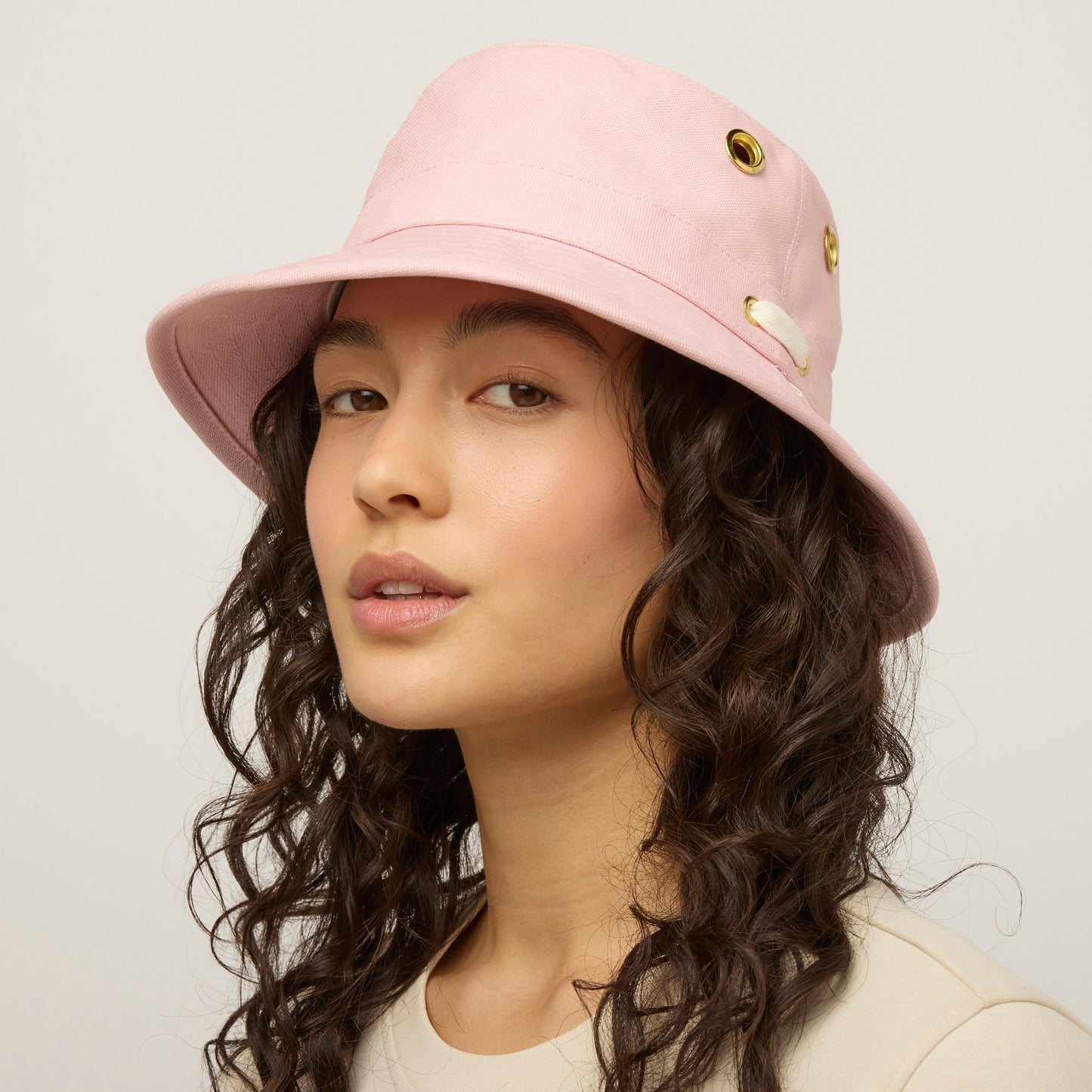 A person with curly hair wears a light pink Tilley T1 Iconic Bucket Hat and a beige top, facing the camera.