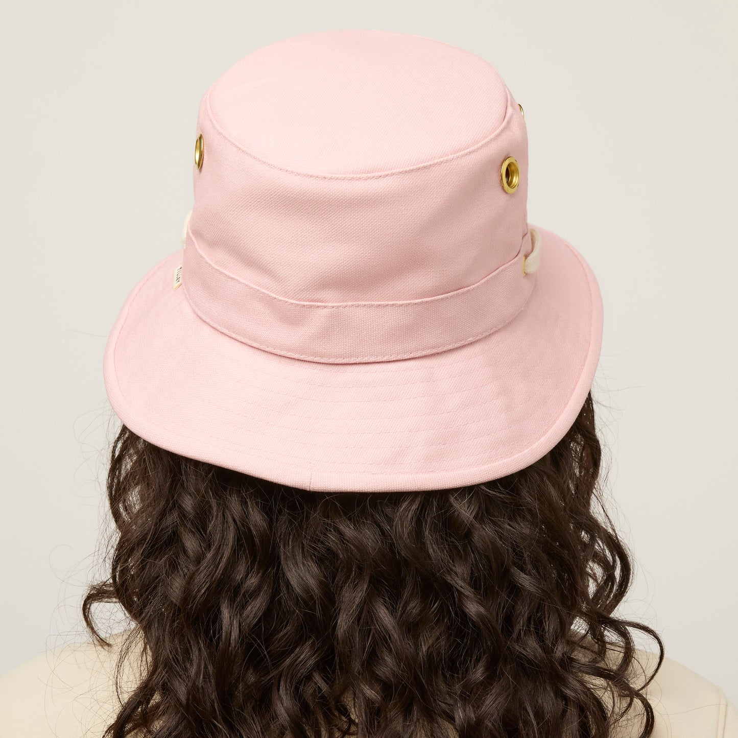 Person with curly brown hair wears a light pink Tilley T1 Iconic Bucket Hat, shown from behind on a neutral background.