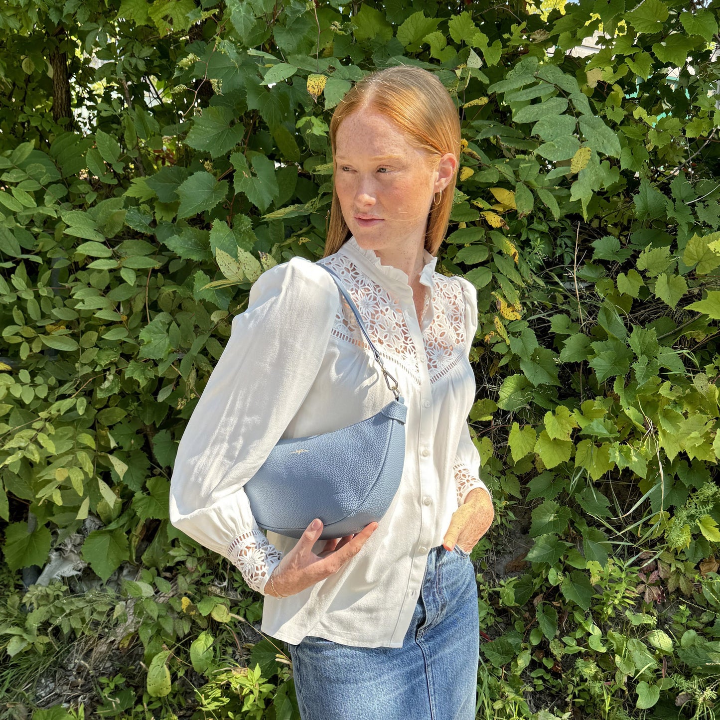 A person holds a blue Espe Crescent Crossbody against lush green leaves.