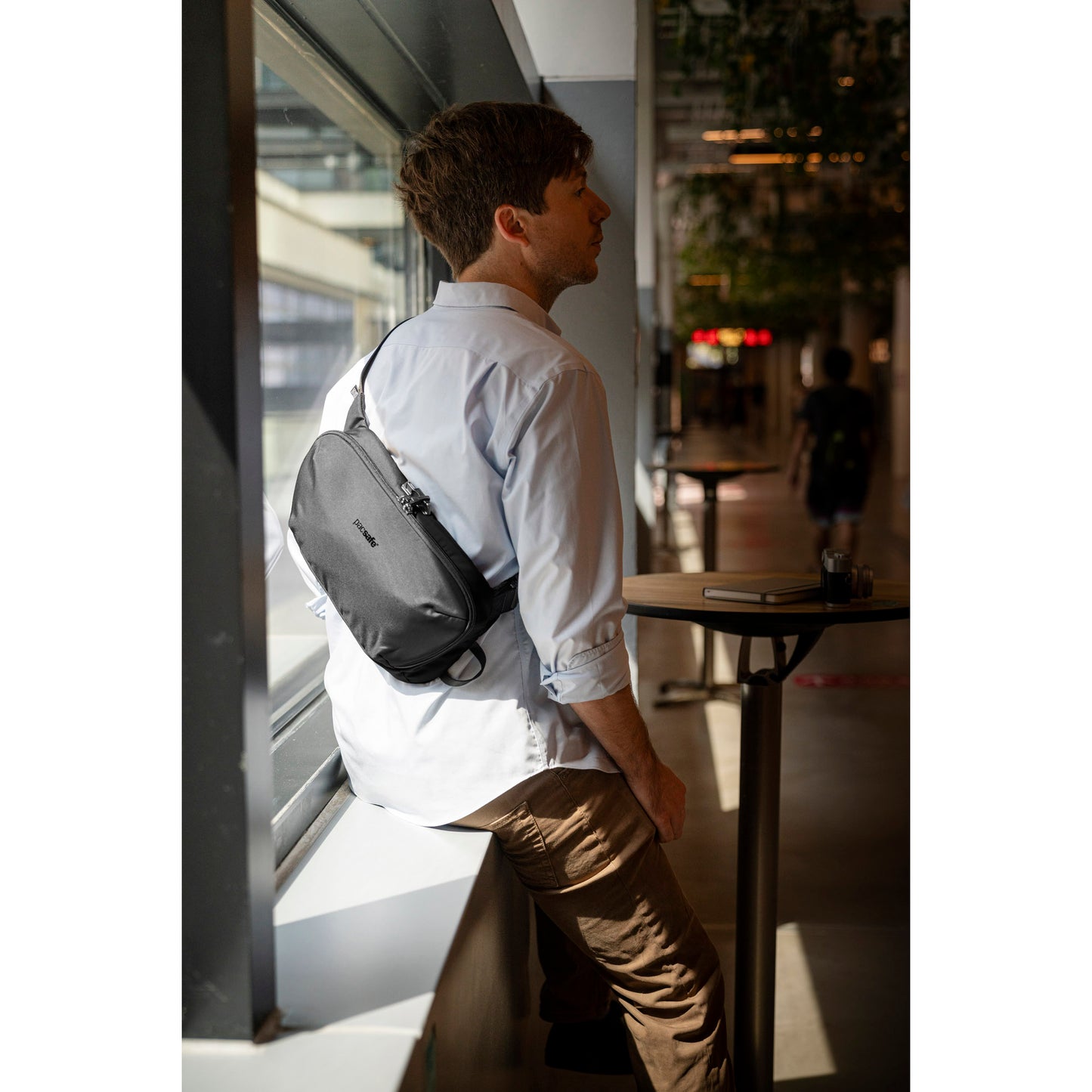 A person in a white shirt and brown pants leans against a window ledge, carrying the Pacsafe® V Anti-theft Urban Sling.