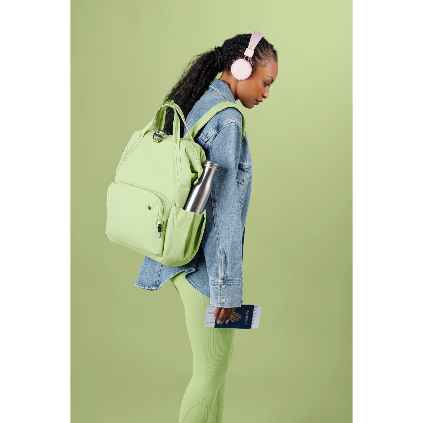 Person in headphones, Pacsafe® CX Anti-theft 17L Backpack & leggings, holding a passport against a green backdrop.