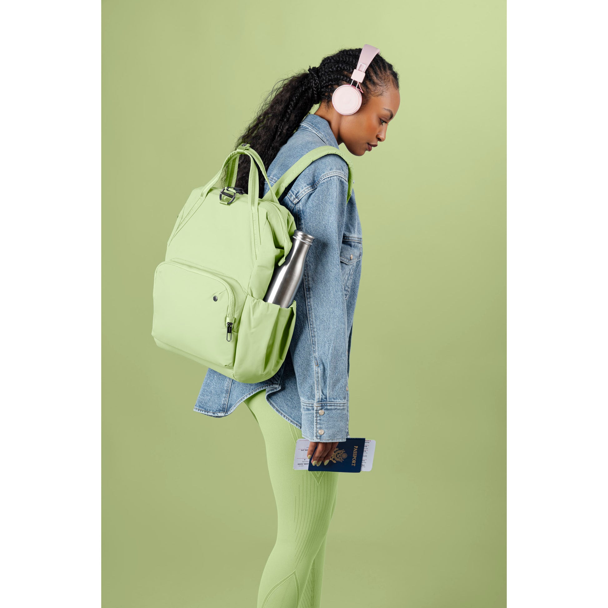 Person in headphones, Pacsafe® CX Anti-theft 17L Backpack & leggings, holding a passport against a green backdrop.
