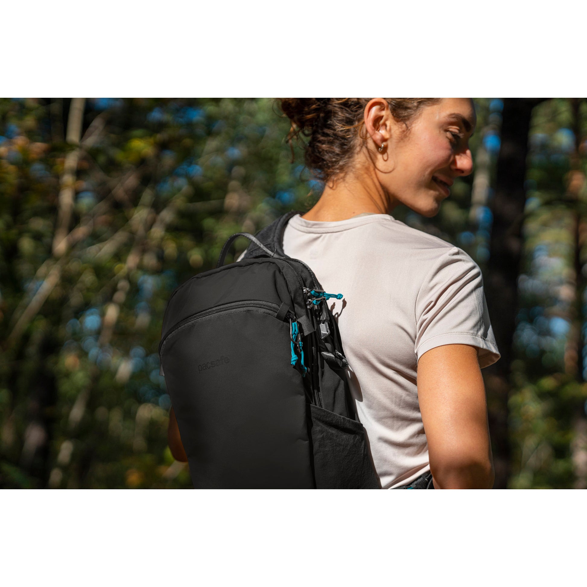 A woman on a forest trail in a beige shirt carrying a Pacsafe® ECO 12L Anti-theft Sling Backpack in black.