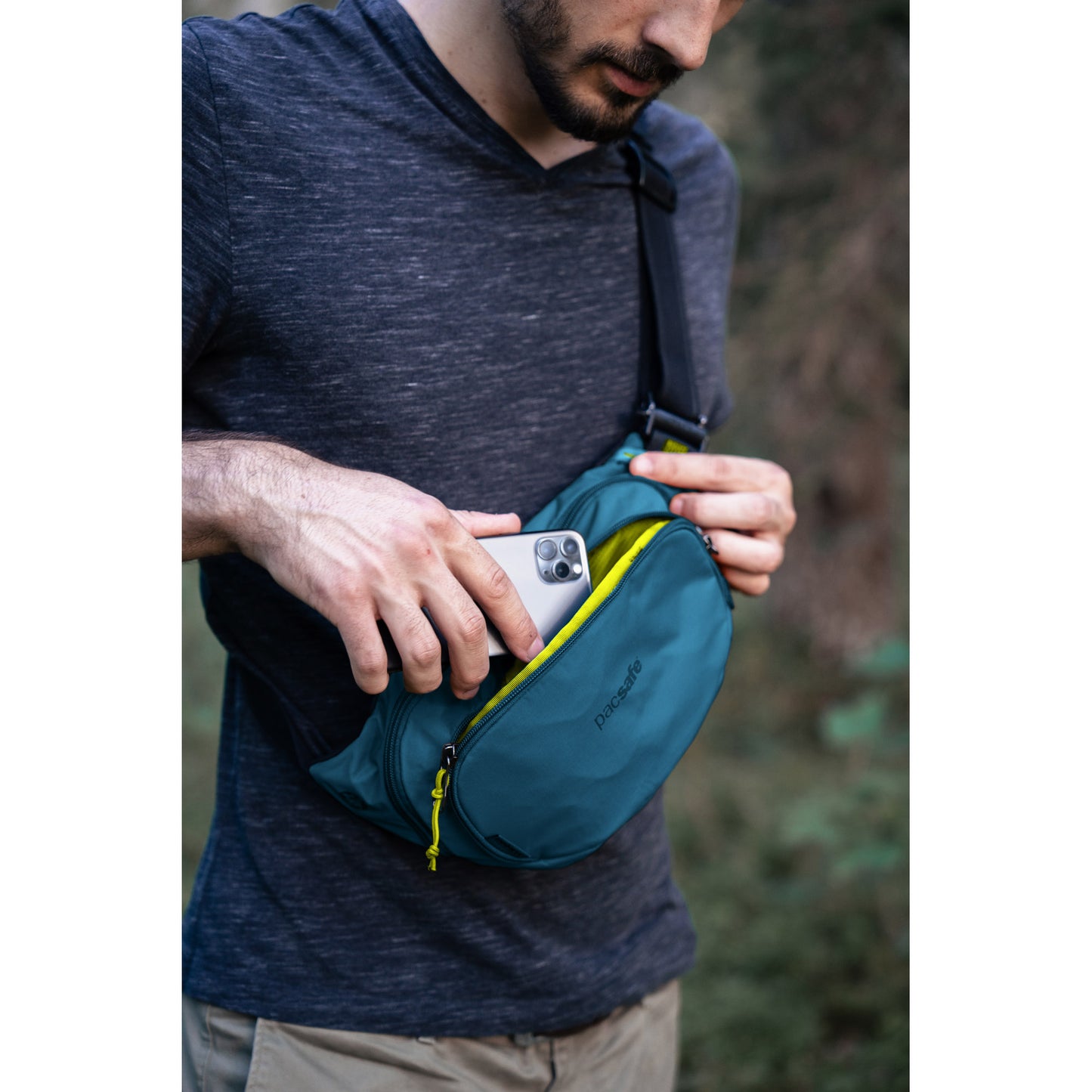 Bearded person puts phone into a Pacsafe® ECO Anti-Theft Waist Pack outdoors, made from ECONYL regenerated nylon.