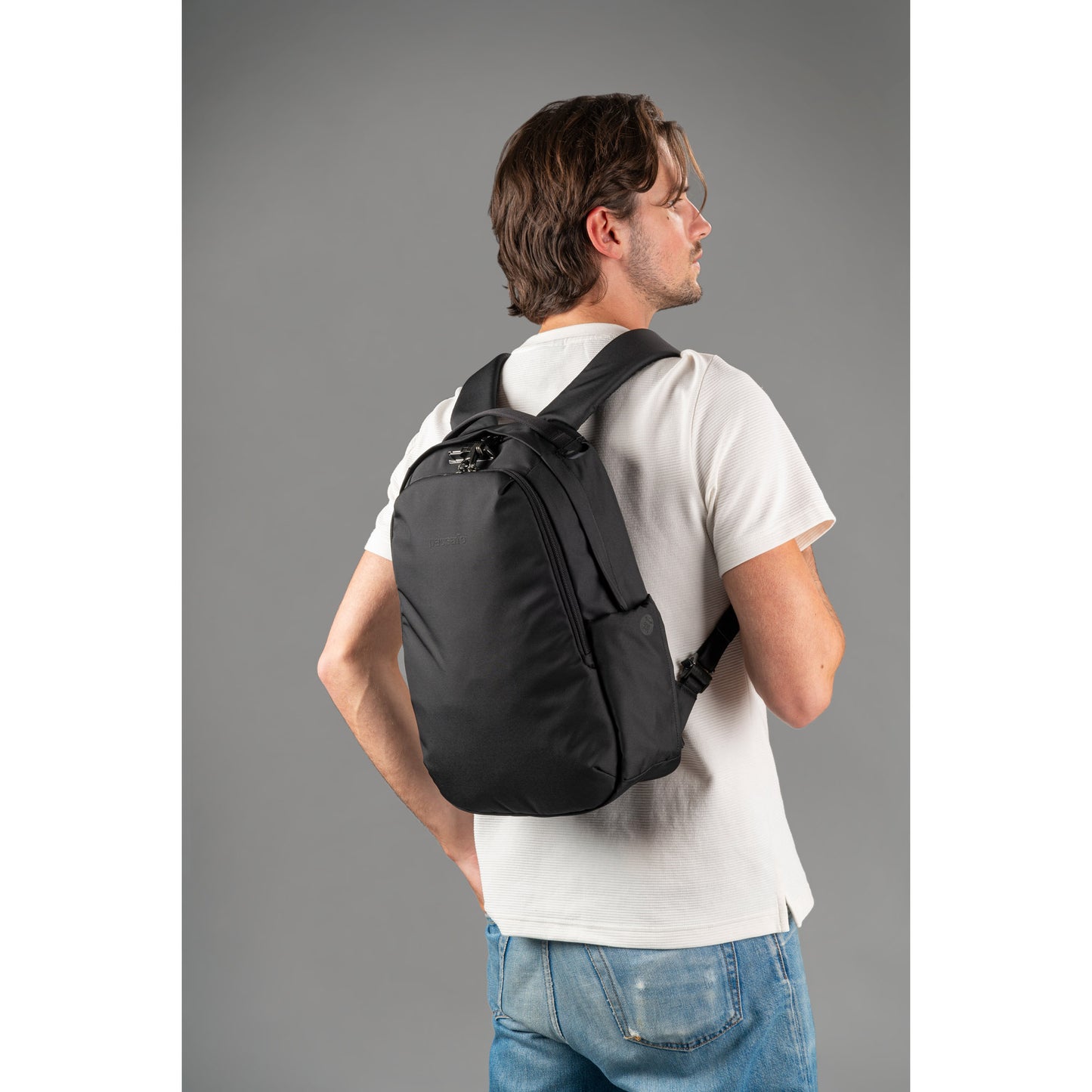 Person with a black Pacsafe® V 16L Anti-theft Backpack, in white t-shirt and jeans, against a gray background.