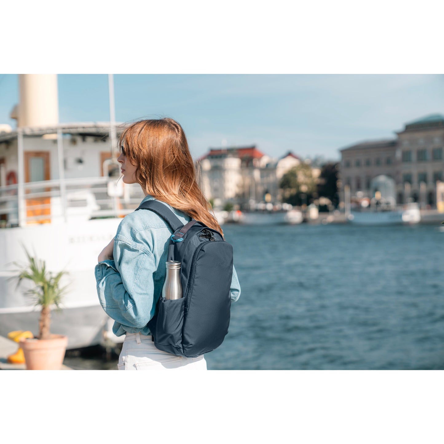 With a Pacsafe® V 16L Anti-theft All-Around Backpack in ocean blue and water bottle, a person stands by a waterfront and buildings.