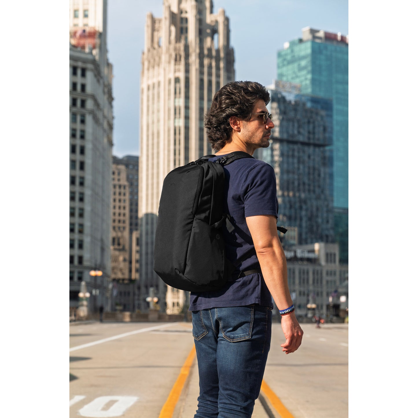 Man with sunglasses and a Pacsafe® V 24L Anti-theft Active Backpack stands on a city street, facing tall buildings.