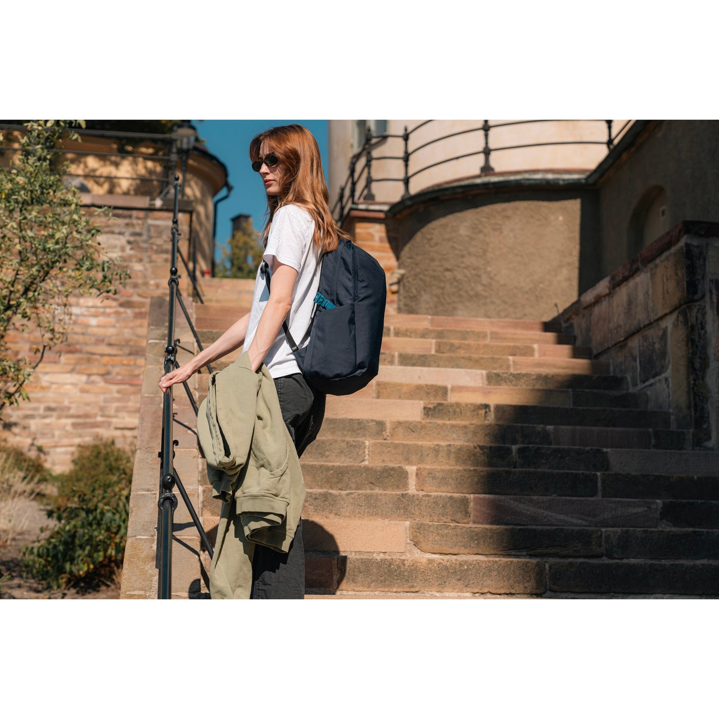 Lifestyle image of woman standing outside on stone steps holding a jacket and wearing the ocean blue Pacsafe® V 24L Anti-theft Active Backpack.