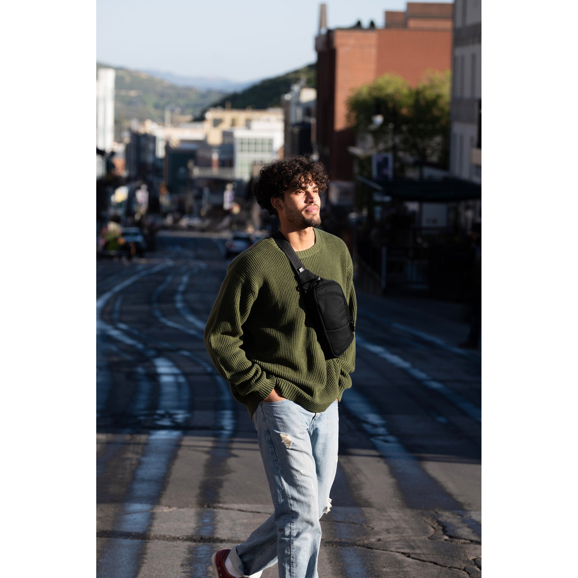 A person in a green sweater and jeans strolls down a sunny street, wearing the Pacsafe® V Anti-theft Action Sling Pack.