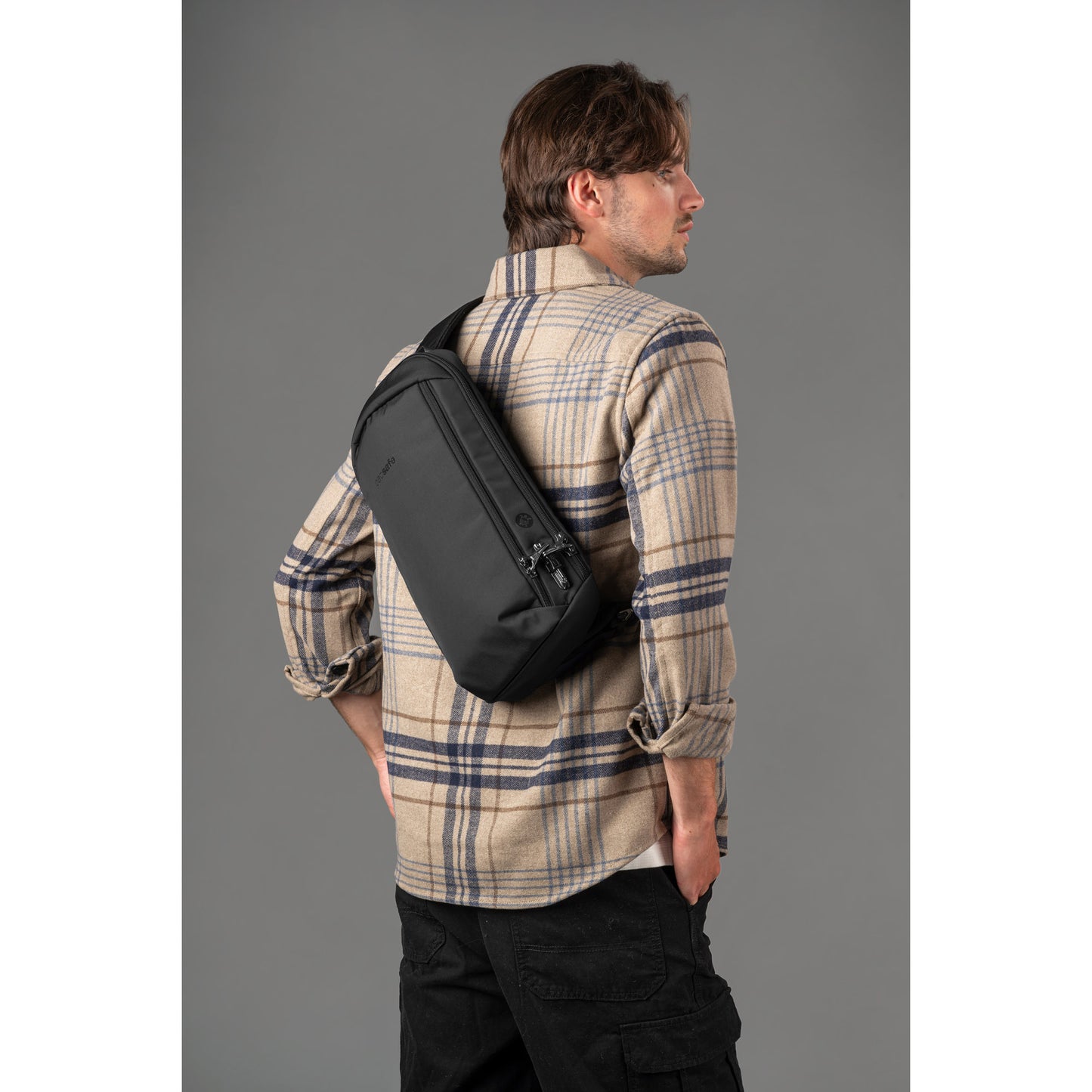 Man in plaid shirt with Pacsafe® V Tech Anti-theft Sling Pack, against a gray background.