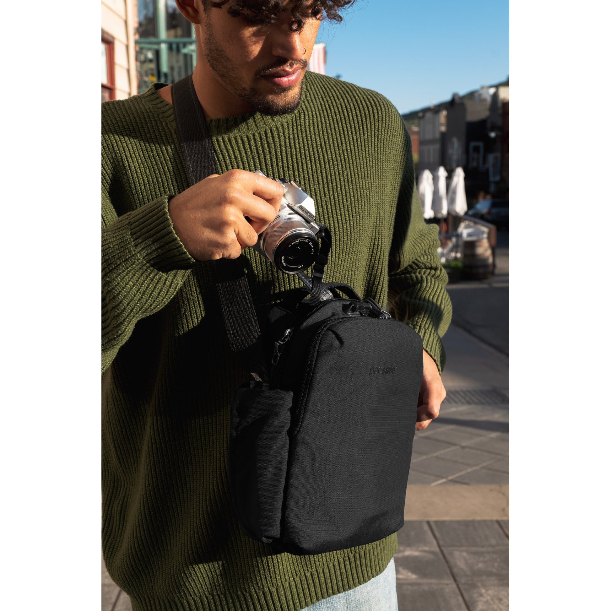 Person in green sweater with Pacsafe® V Anti-theft Tour Crossbody, holding a camera on a city street.