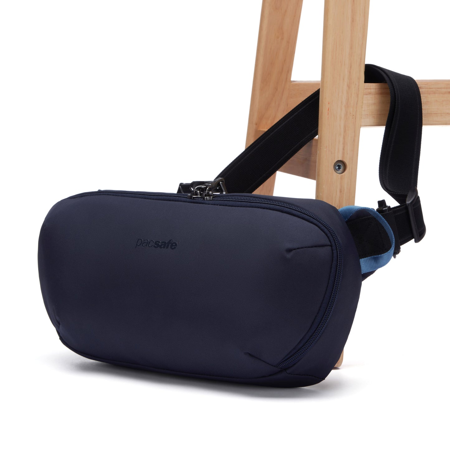 The Pacsafe® V Anti-theft Urban Sling in ocean blue, made from recycled materials, hangs on a chair with black straps.