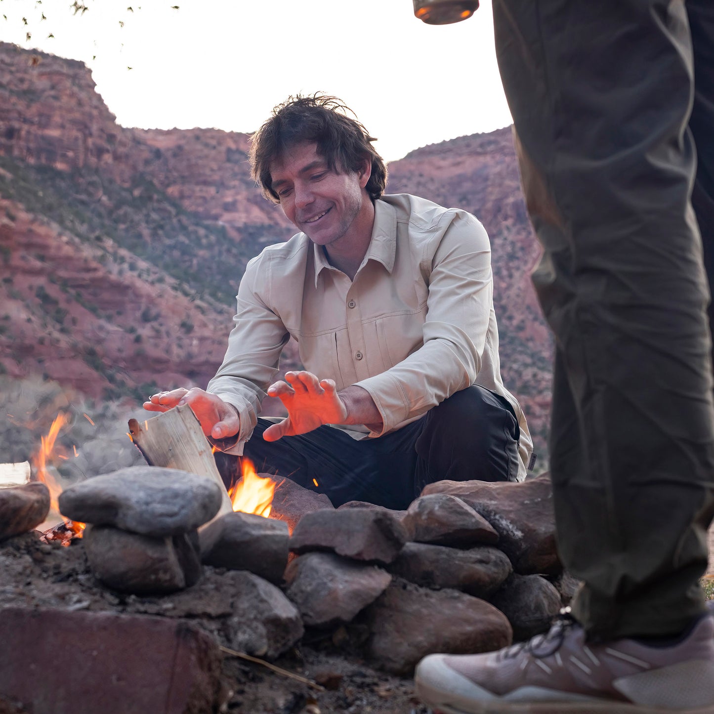Lifestyle image of man wearing Royal Robbins Expedition III long sleeve shirt while sitting in front of a campfire with mountains in the background.