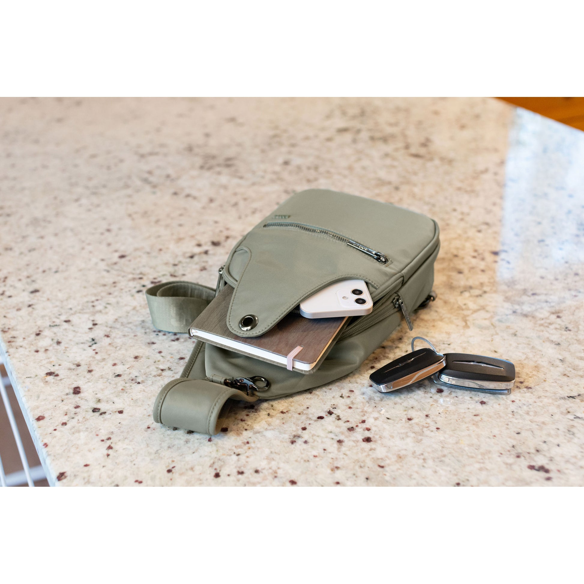 Lifestyle image of the Roots Travel Sling Bag in sage colour, laying on a marble countertop with a notebook and phone sticking out and car keys beside it.