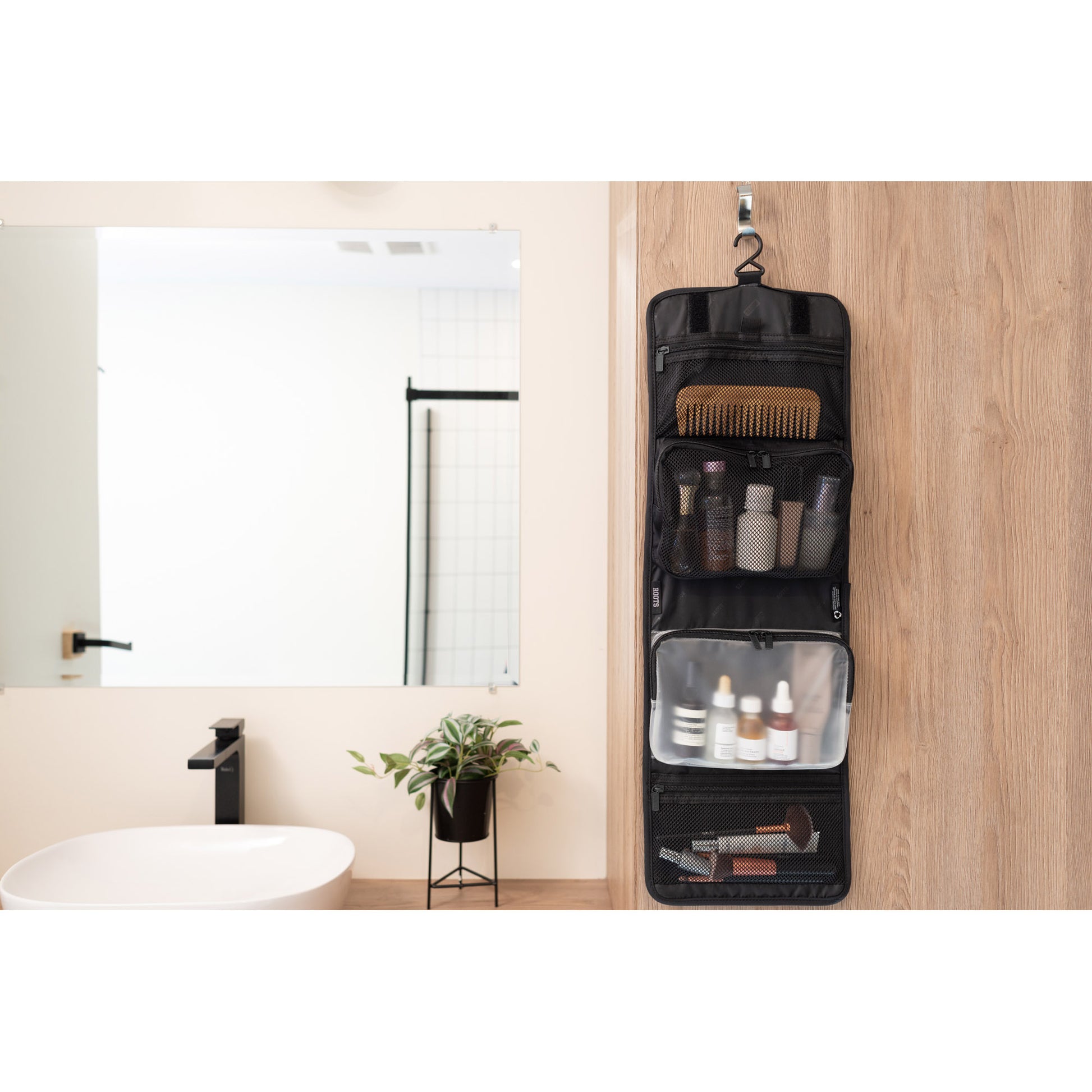 Lifestyle image of the Roots Travel Toiletry Bag in black filled with bottles, make-up and a comb, hanging open on a hook in a bathroom.