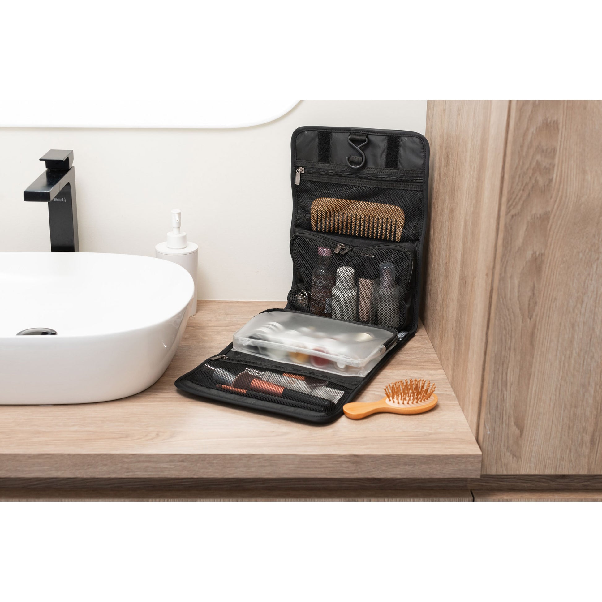 Lifestyle image of Roots Travel Toiletry Bag in black filled with small bottles, make-up and hair accessories, open and propped up beside a sink on the counter.