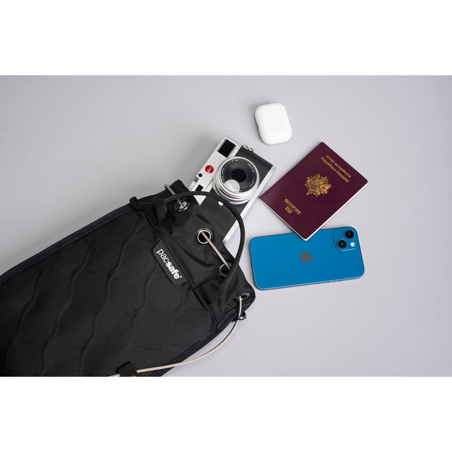 Pacsafe® Travelsafe® 3L holding a camera, phone, passport, and earbuds on a gray surface.