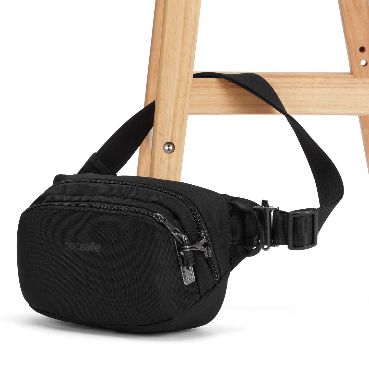 The Pacsafe® V Anti-theft Hip Pack, a black recycled polyester crossbody bag with lockable zips, secured to a wooden stool leg.