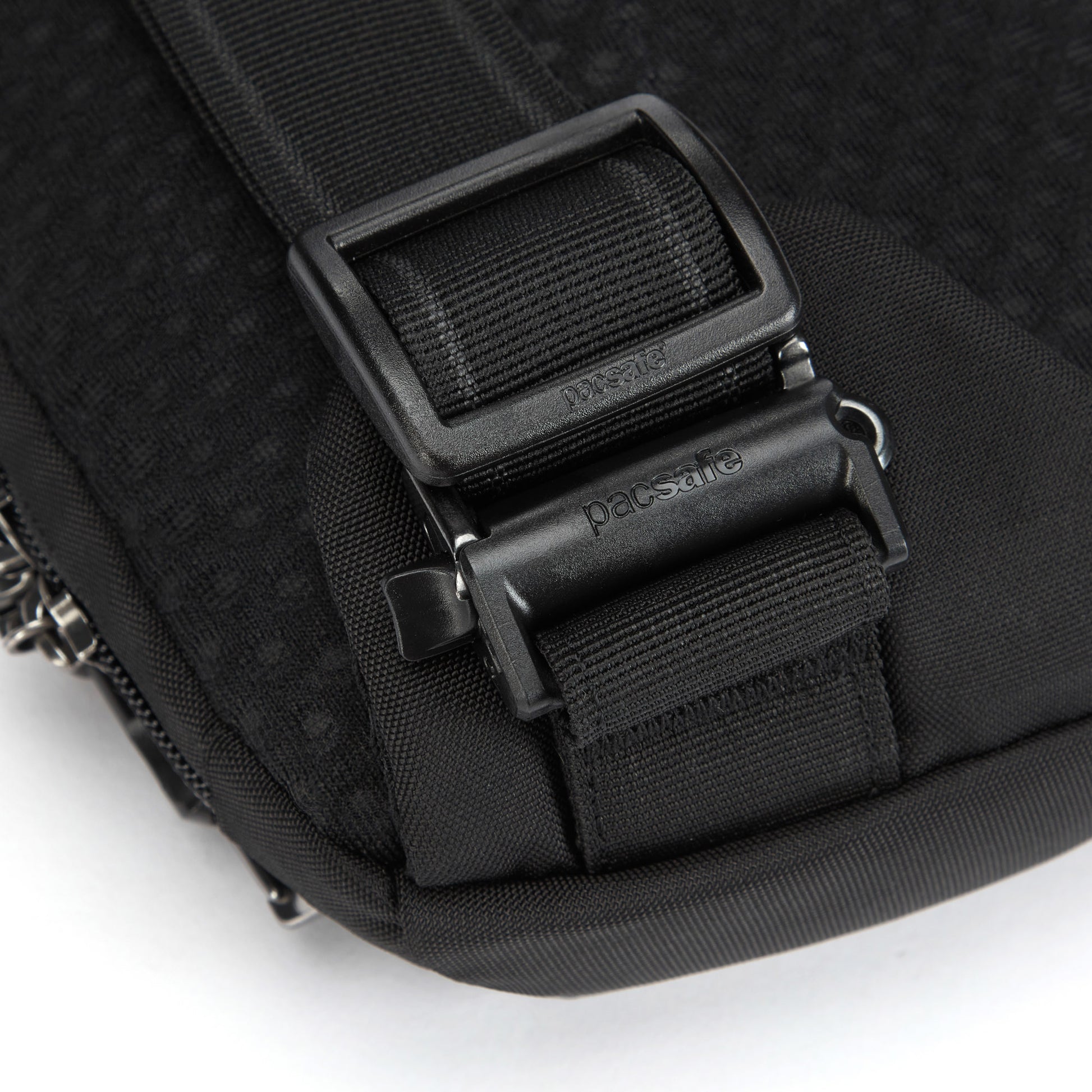 Close-up of Pacsafe® V Sling Pack with black strap, plastic buckle, and metal clip on textured fabric.