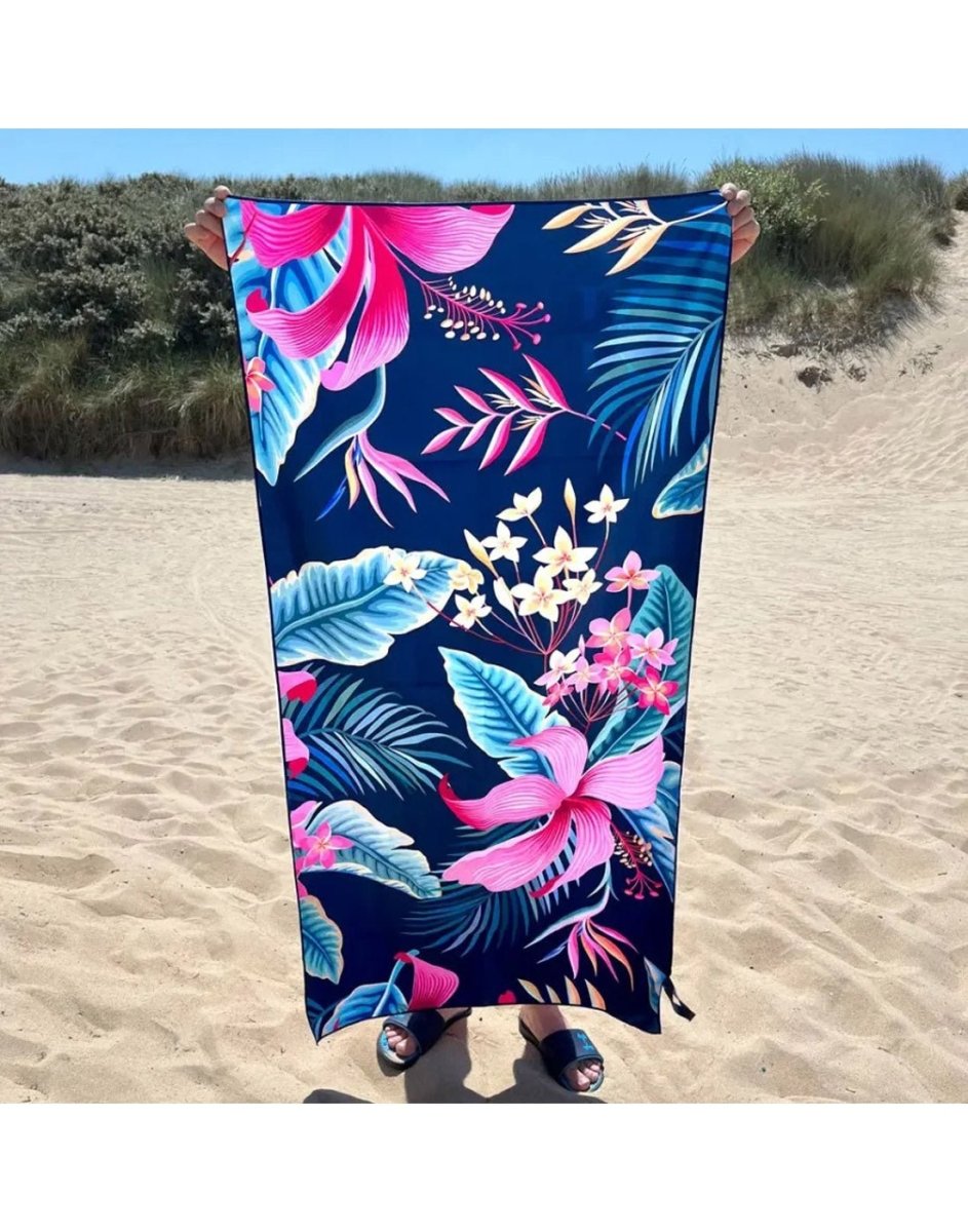 Person holding the K5 Microfibre Beach Towel with pink and white tropical flowers on a sandy beach.