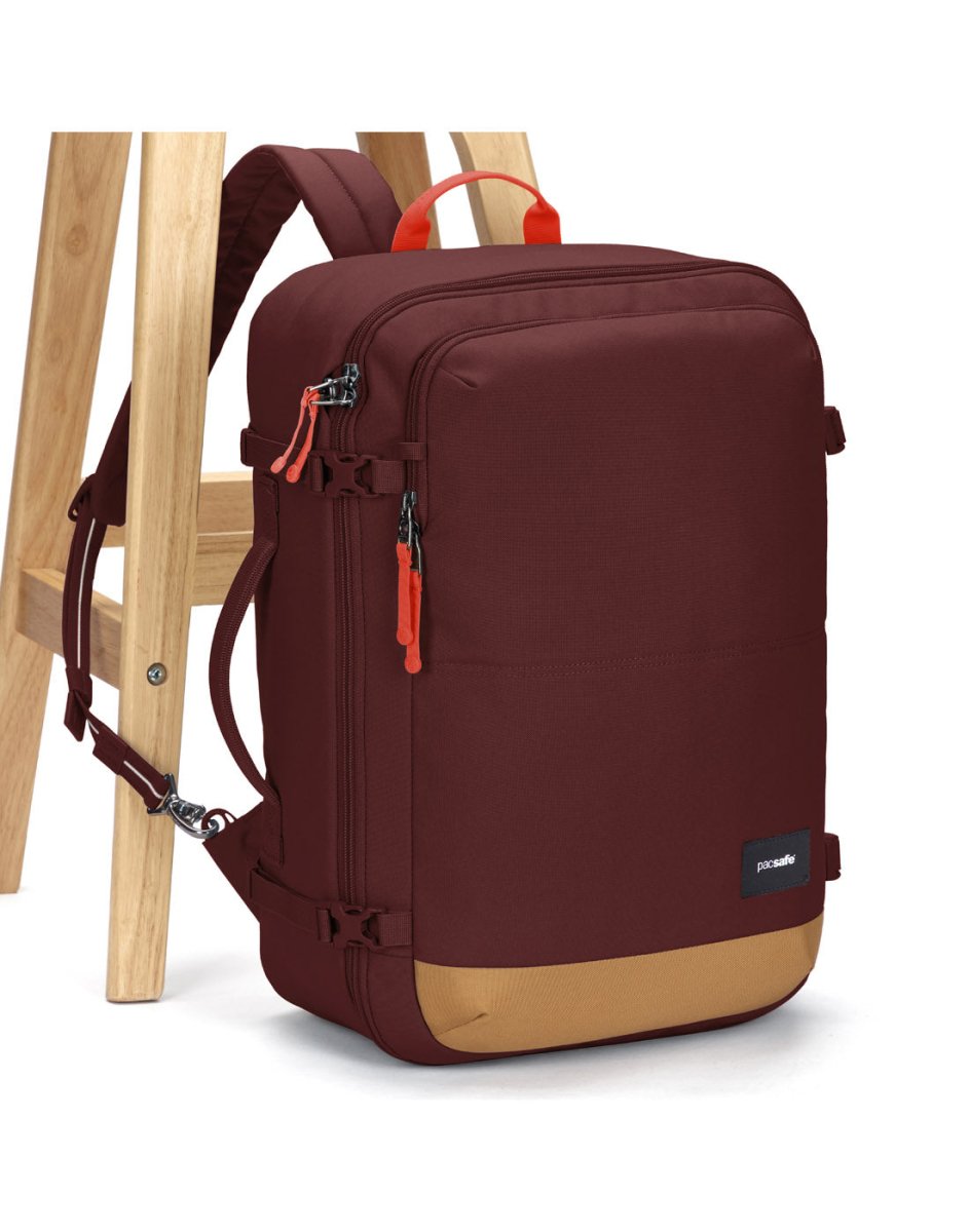 The Pacsafe® GO 34L Anti-theft Carry-on Backpack in maroon/tan with orange zippers rests against a stool.