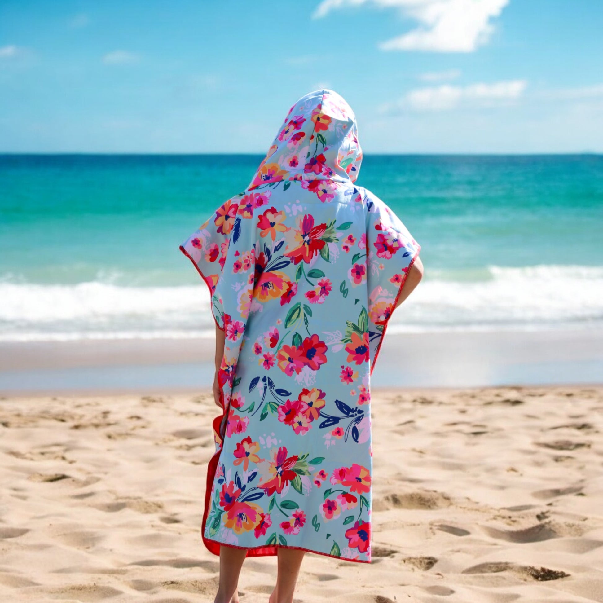 A child wearing the K5 Microfibre Poncho stands on a sandy beach under a partly cloudy sky.