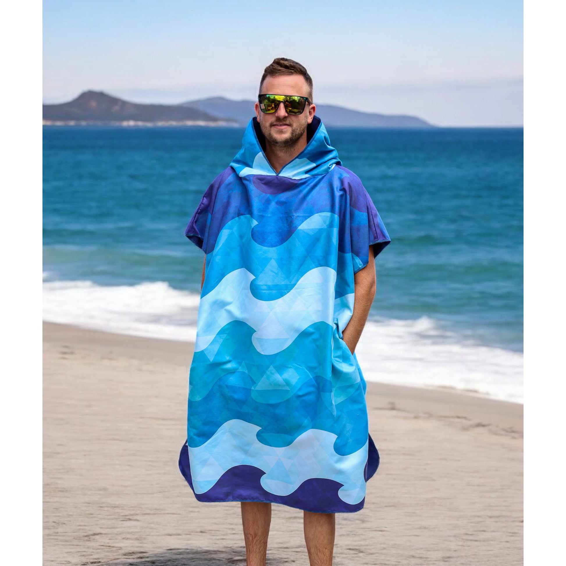A man in a K5 Microfibre Poncho - Adult stands on a sandy beach with the ocean in the background.