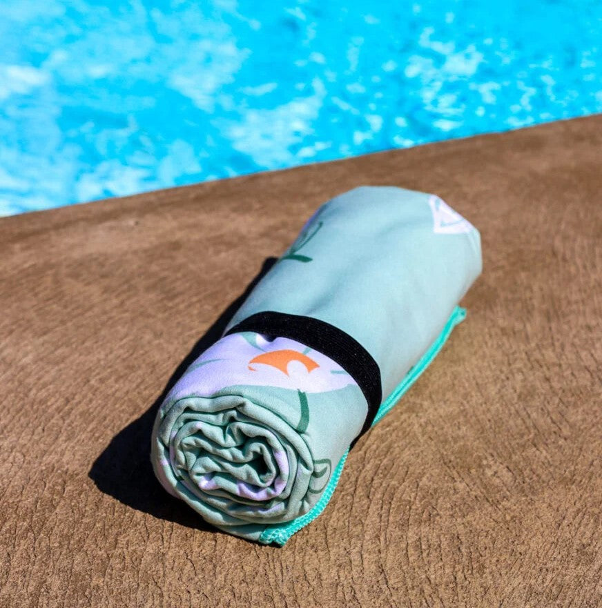 The K5 Microfibre Beach Towel with a black strap rests poolside by the shimmering blue water.