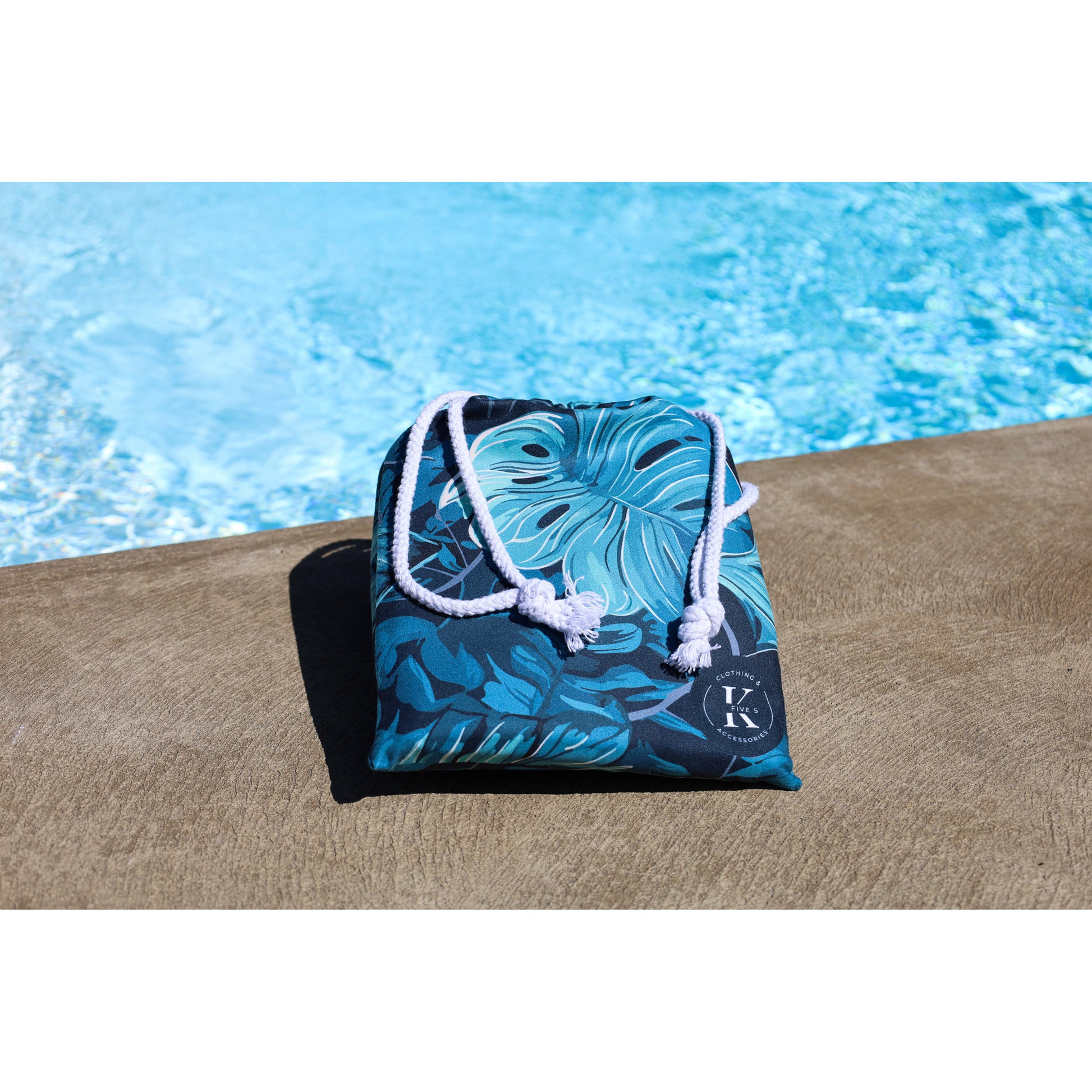 A folded K5 XXL Microfibre Beach Towel in blue with white rope straps sits poolside by clear water.
