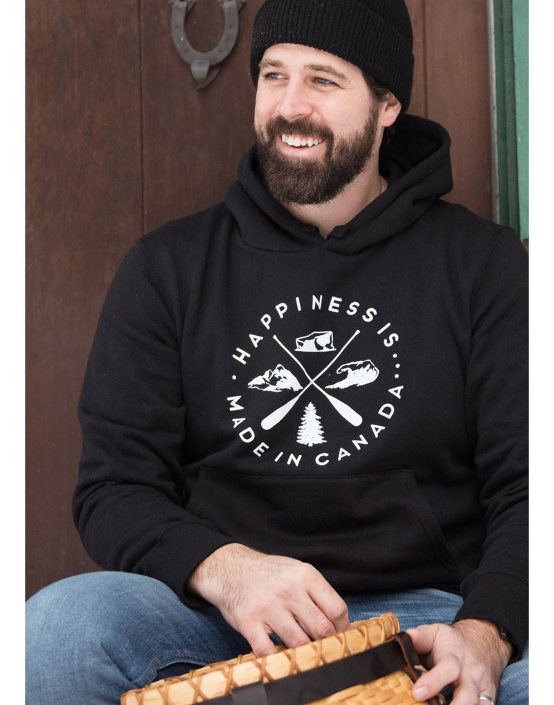 Lifestyle image of a man wearing blue jeans, black toque and Happiness Is...Unisex Crest Hoodie in black, sitting infront of a wooden door