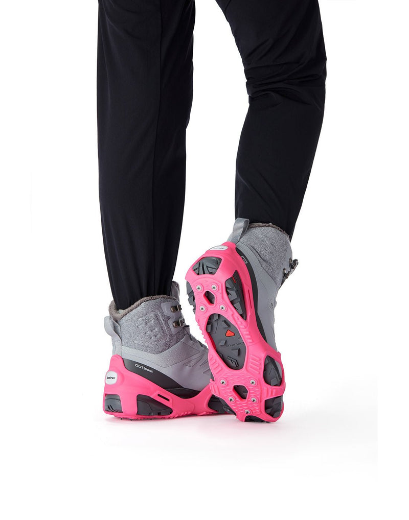 Legs from knee down wearing black pants and grey hiking boots with the Icetrax V3 Tungsten Ice Cleats in pink on them