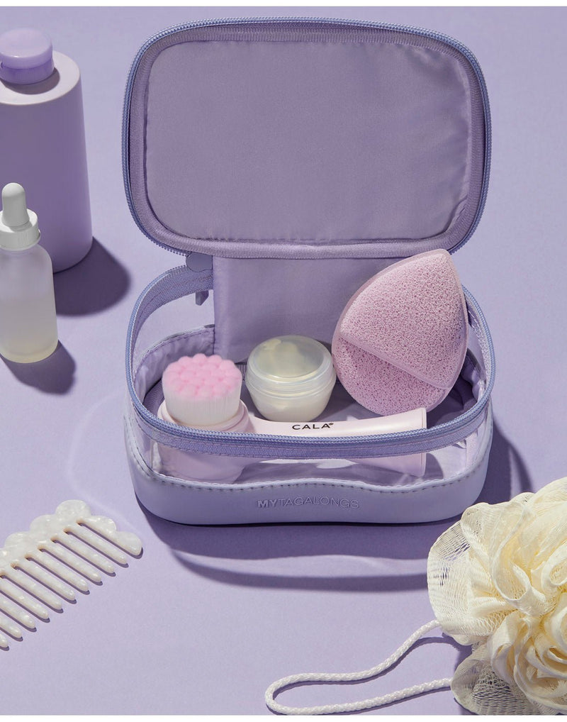 Purple product spread - light lilac mini train case, popped open on a purple surface with make-up sponges and small containers inside.  There is a white comb, white shower puff, clear dropper bottle and purple bottle beside the open case.
