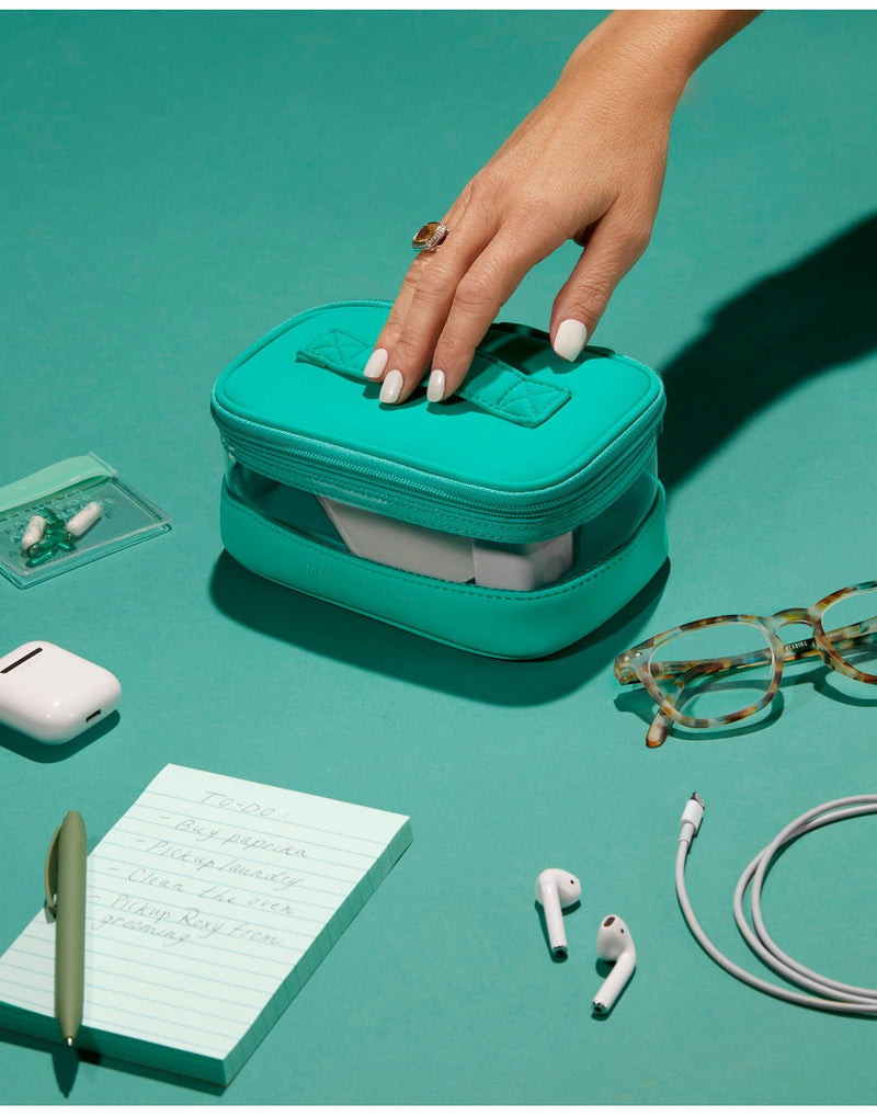Clover product spread - clover mini train case on a teal colour surface with a women's hand on top, reaching for it. There is a note pad with green pen, white ear buds, cord and case, as well as green tortoise shell glasses and small clear zip pouch with pills spread around the case.
