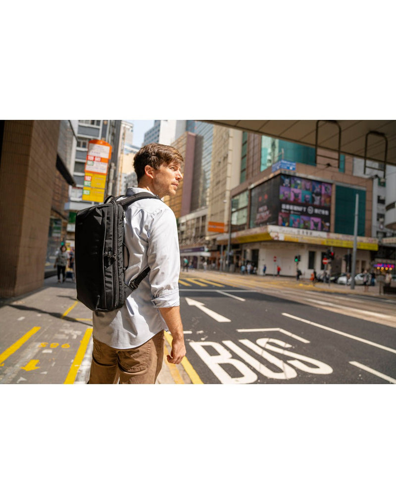 Lifestyle image of a man wearing a white shirt and tan pants with the Pacsafe Metrosafe X Anti-Theft 16-Inch Commuter Backpack in black, ready to cross a busy city street
