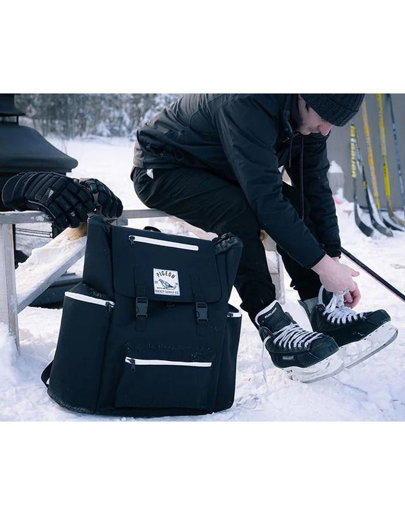 Man outdoors in winter sitting on a bench putting on hockey skates with the Pigeon Pack on the snow beside him