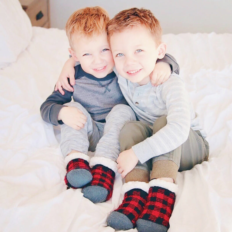 Two young children sitting on a bed with arms around each other, both wearing Pudus Kids Classic Slipper Socks in Lumberjack Red 