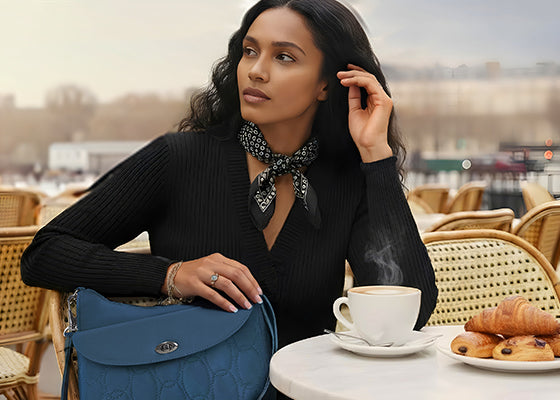 Woman sitting on an outside cafe nicely dressed with a blue purse and coffee and croissants on the table.