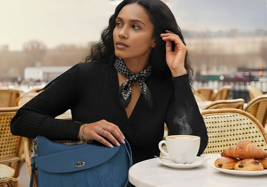 Woman sitting on an outside cafe nicely dressed with a blue purse and coffee and croissants on the table.