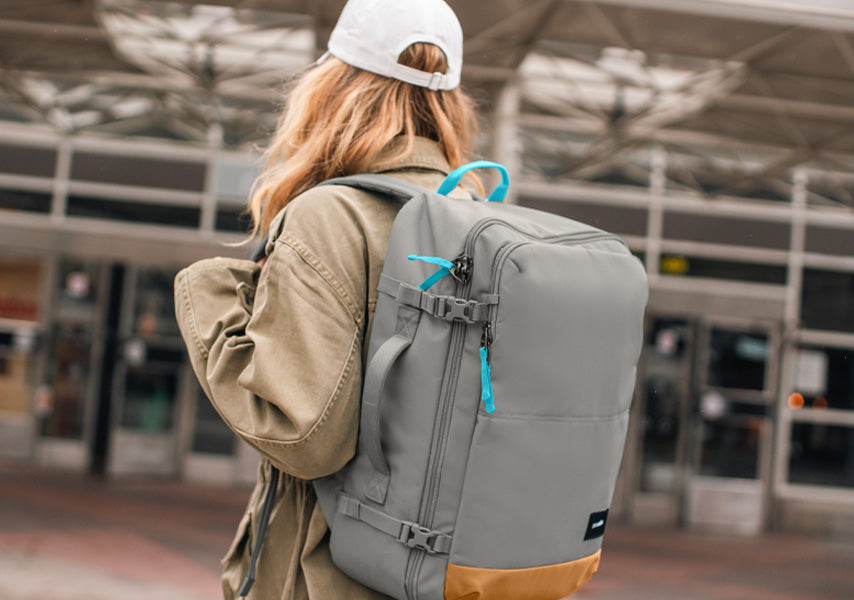 Woman walking towards doors of a train station carrying a Pacsafe® GO 44L Anti-theft Carry-on Backpack in stone grey.