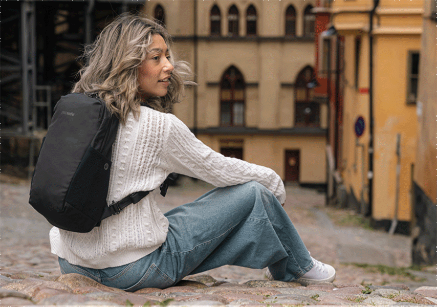 Man with black backpack, getting into a white old offroad car. Woman sitting on the higher side of the street, looking to the side with colourful tall building around in an old style. 