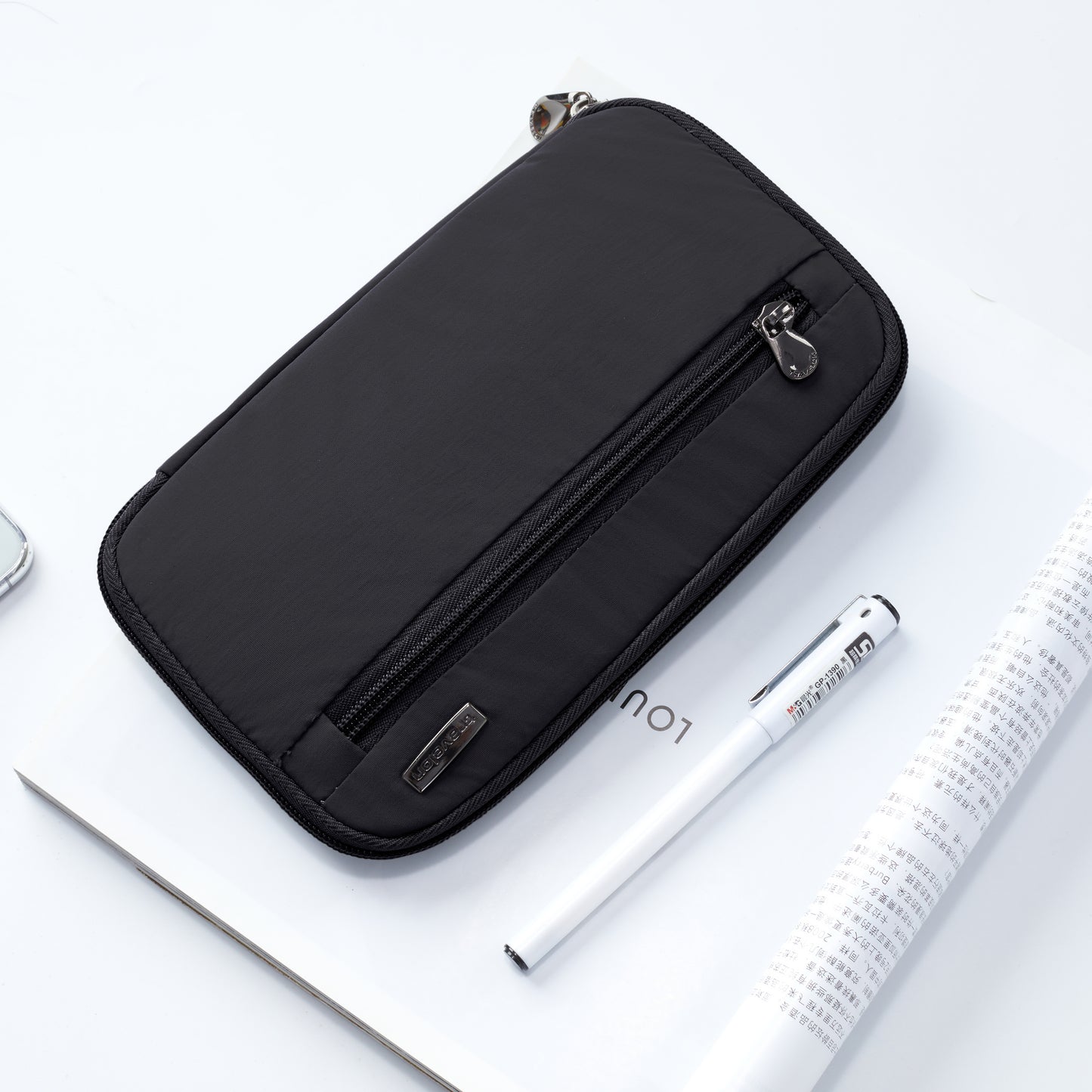 The Travelon RFID Blocking Zip Passport Wallet in black sits next to a white pen and rolled magazine on a white surface.