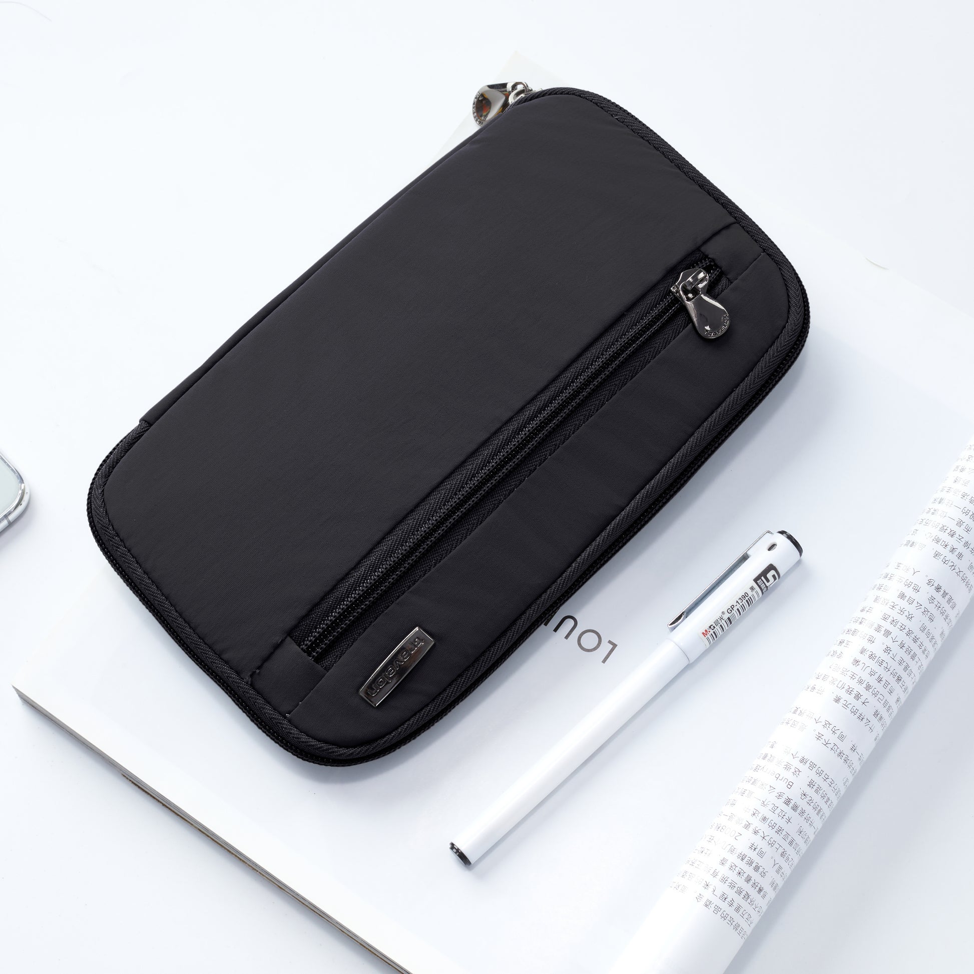 The Travelon RFID Blocking Zip Passport Wallet in black sits next to a white pen and rolled magazine on a white surface.