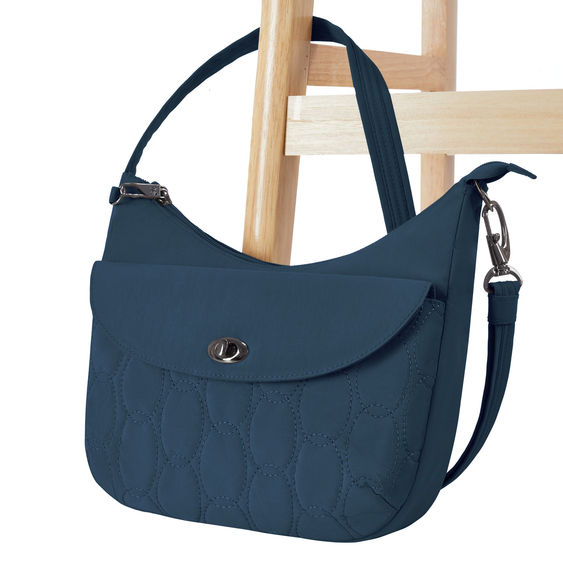 The Travelon Runways Anti-theft Medium Crossbody in blue hangs on a wooden chair, flaunting chic quilted style.