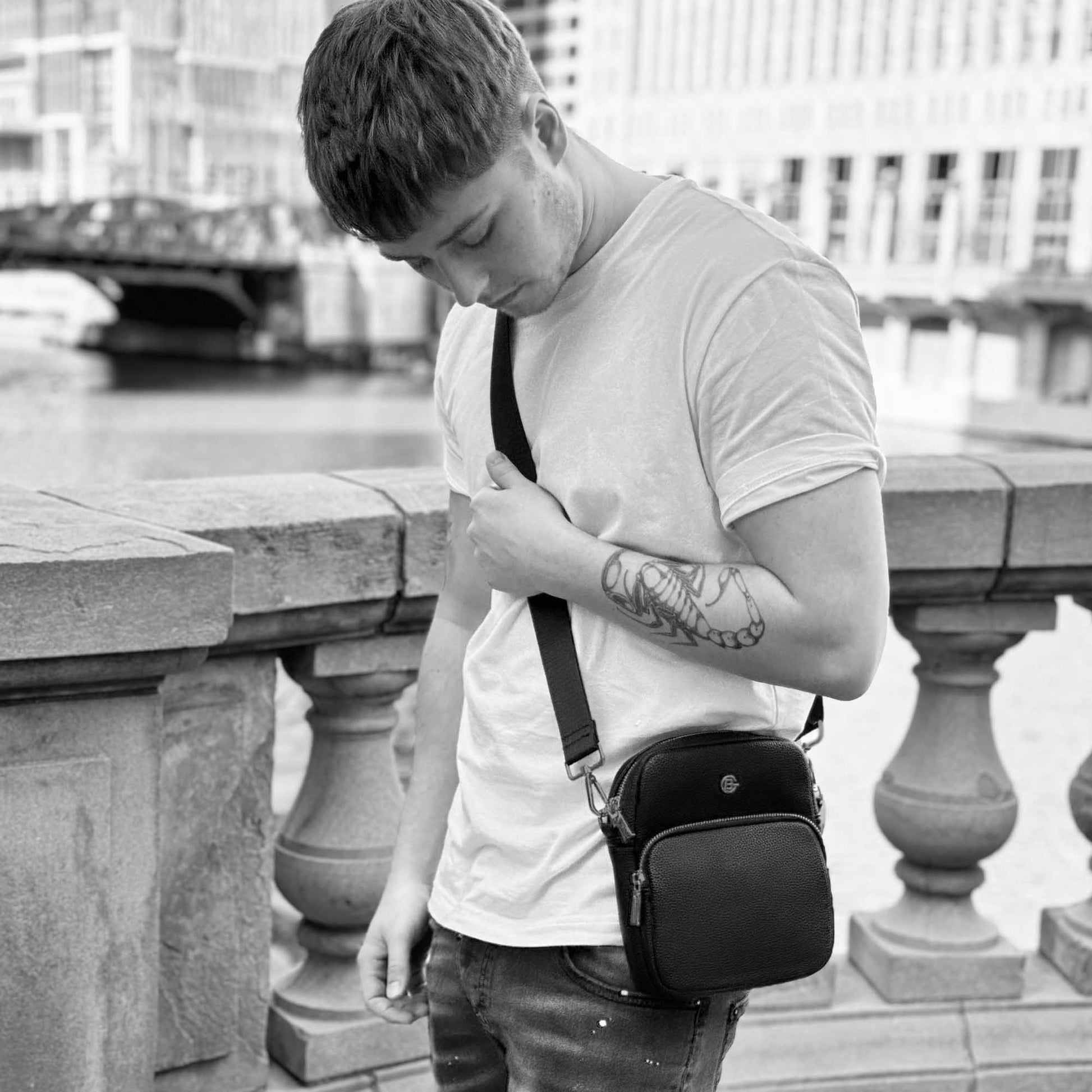 Black and white lifestyle image of man standing on a bridge wearing the black Espe Art Crossbody